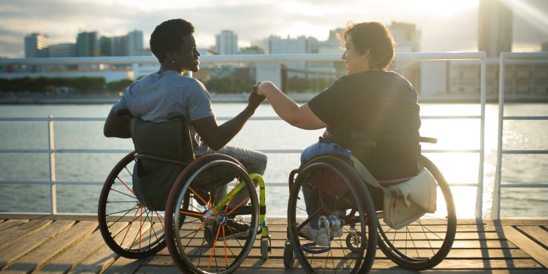 Fond girlfriend and boyfriend spending day together. African American man and Caucasian woman in wheelchairs, holding hands, looking at sunset. Love, affection, happiness concept